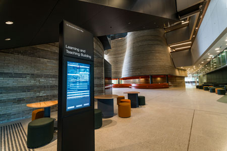 Monash University foyer with a Learning and Teaching Building digital sign, stools and tables