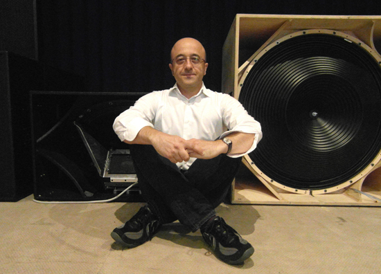 Massimo Minardi seated on the floor between a loudspeaker cabinet and a large subwoofer cone