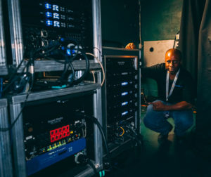 Man with a lanyard crouching beside touring racks of amplifiers with blue front displays