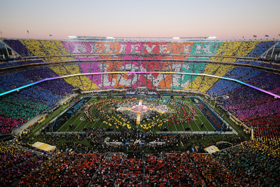 Super Bowl 50 halftime show seen from above, with a crowd card display spelling BELIEVE IN LOVE