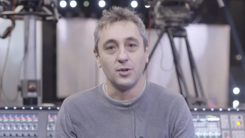Massimo Faggioni speaking to camera in front of a mixing console and stage lights