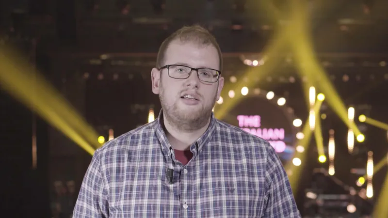 Robin Conway speaking to camera in front of a stage with yellow light beams